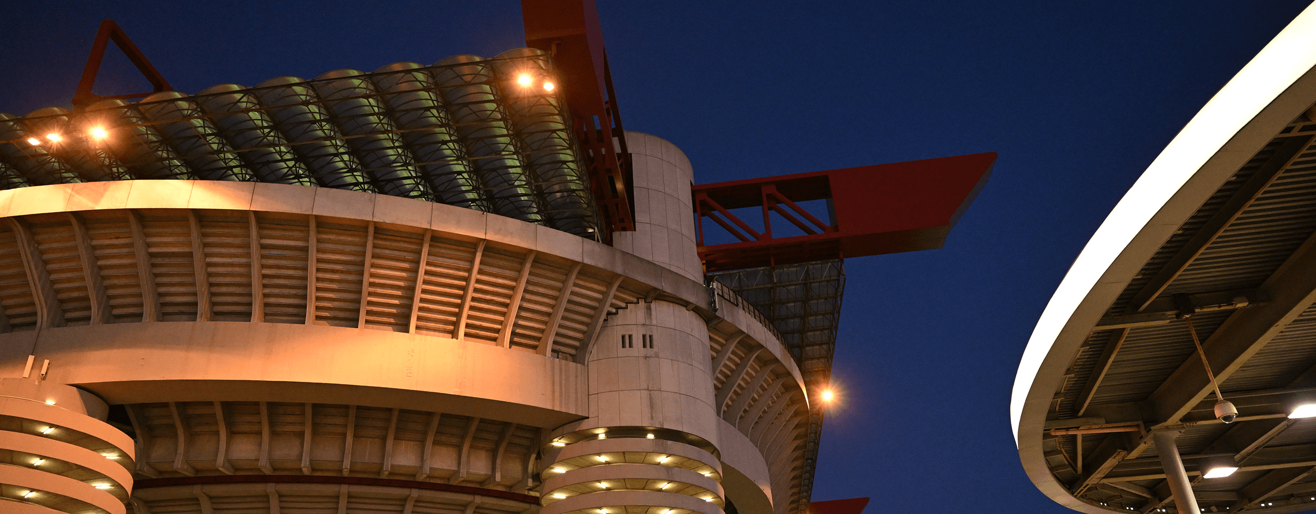 Evening exterior view of San Siro lit up, showing the towers and red structures in detail